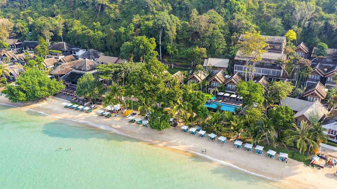 Luftfoto af en strand med liggestole og parasoller samt bygninger omkranset af grønne områder. (AI-genereret tekst)