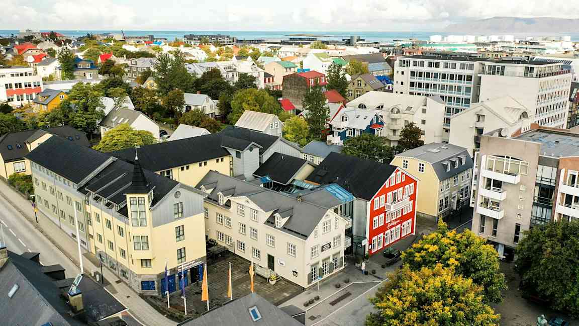 En byudsigt med farverige bygninger, træer og en vej, samt baggrund med hav og bjerge. (AI-genereret tekst)