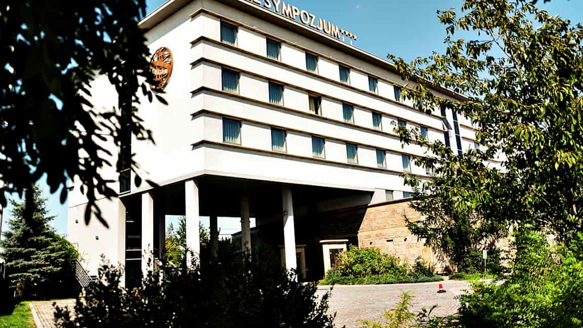 Bygning med flere etager, mærket med teksten 'Hotel Sympozjum', omgivet af træer og vegetation. (AI-genereret tekst)