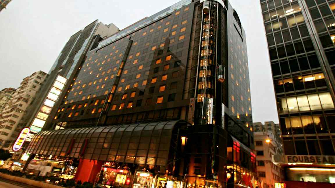 En oplyst fleretagers bygning i et bymiljø med glasfacade og gadeliv i forgrunden. (AI-genereret tekst)