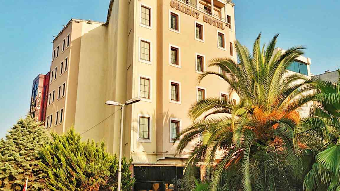 En hotelbygning med flere etager. Der er palmer og grønne træer foran bygningen. (AI-genereret tekst)