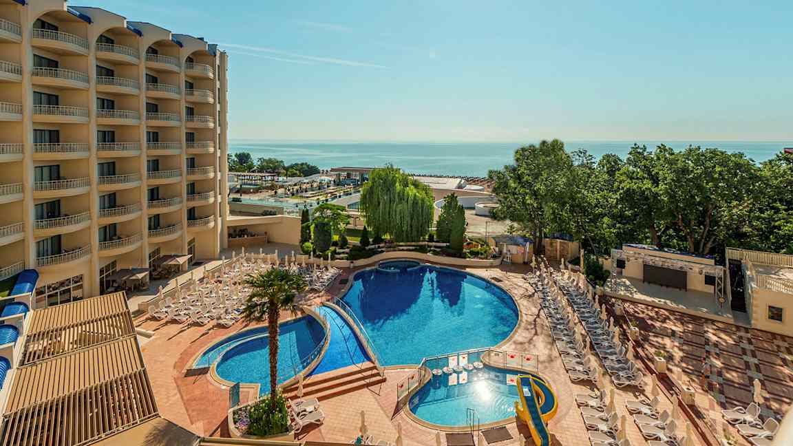 Billede af et hotel med en stor udendørs pool, liggestole og omkringliggende vegetation. Bygningen har flere etager med altaner. (AI-genereret tekst)