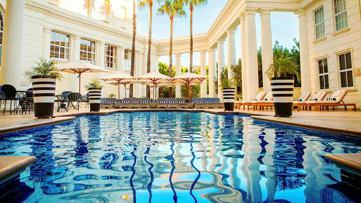 Poolområde med glinsende blåt vand, liggestole og parasoller omgivet af søjler og palmer. (AI-genereret tekst)