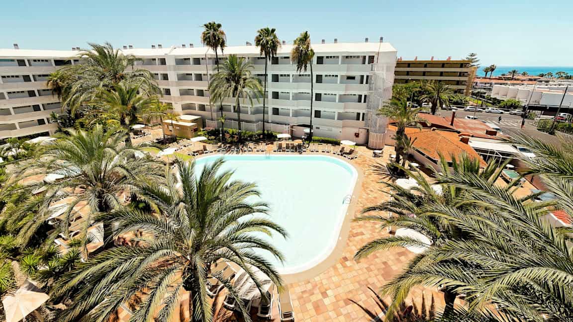 En udendørs pool omgivet af liggestole og palmer. I baggrunden ses et hotel med hvid facade. (AI-genereret tekst)