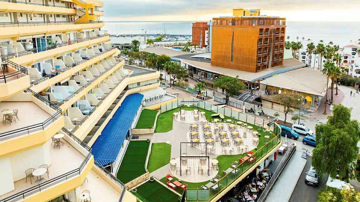 Hotel med gule facader og altaner, en terrasse med borde og stole omgivet af grønne områder samt en udsigt over byen og havet i baggrunden. (AI-genereret tekst)