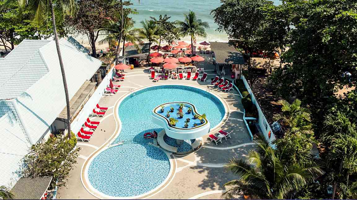 Luftfoto af en pool på et feriested med liggestole og parasoller, omgivet af tropiske træer og tæt på havet. (AI-genereret tekst)