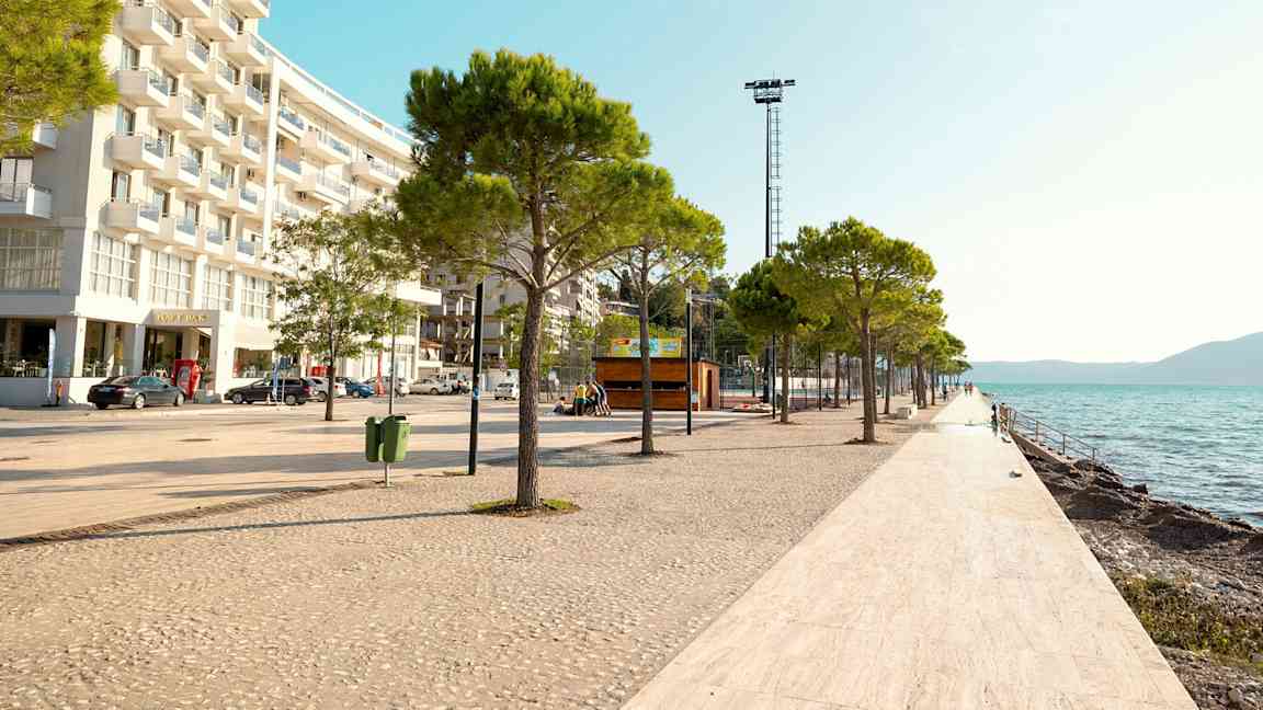 En strandpromenade med træer og hvidflisebelagt gulv langs vandet, med en moderne bygning til venstre. (AI-genereret tekst)