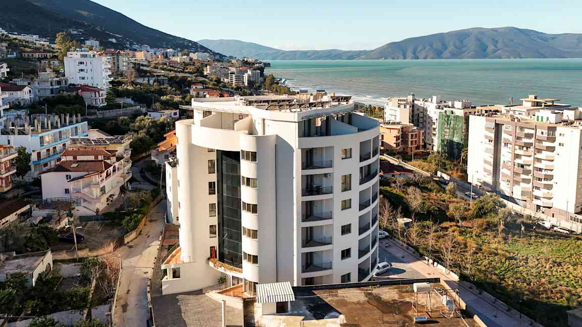 En høj bygning tæt på havet, omgivet af bygninger og grønt. (AI-genereret tekst)