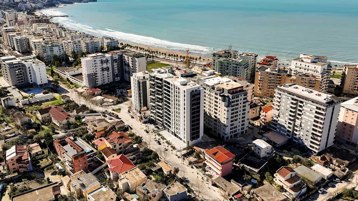 Billedet viser en by med højhuse og boliger tæt på en kystlinje med sandstrand og palmer. (AI-genereret tekst)