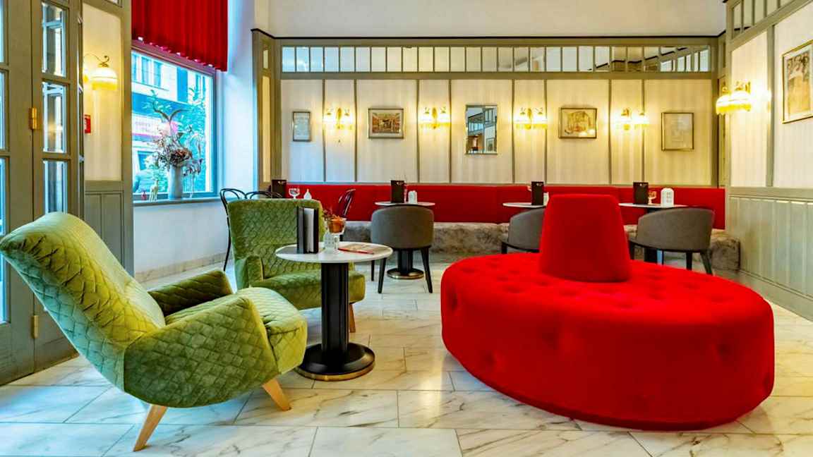 En hotellobby med siddeområde, inklusive grønne lænestole, røde og grå sidemøbler og marmorgulv.  (AI-genereret tekst)