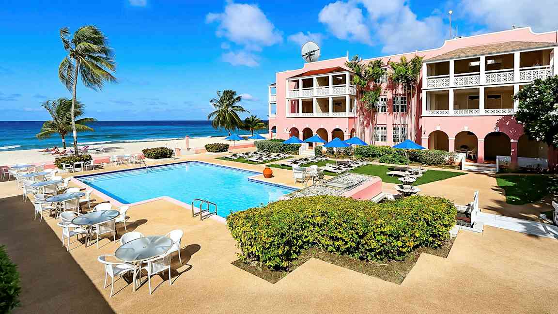 Billede af en solrig ferieby med en udendørs pool, omkringliggende terrasser og et lyserødt hotel med altaner, beliggende ved havet. (AI-genereret tekst)