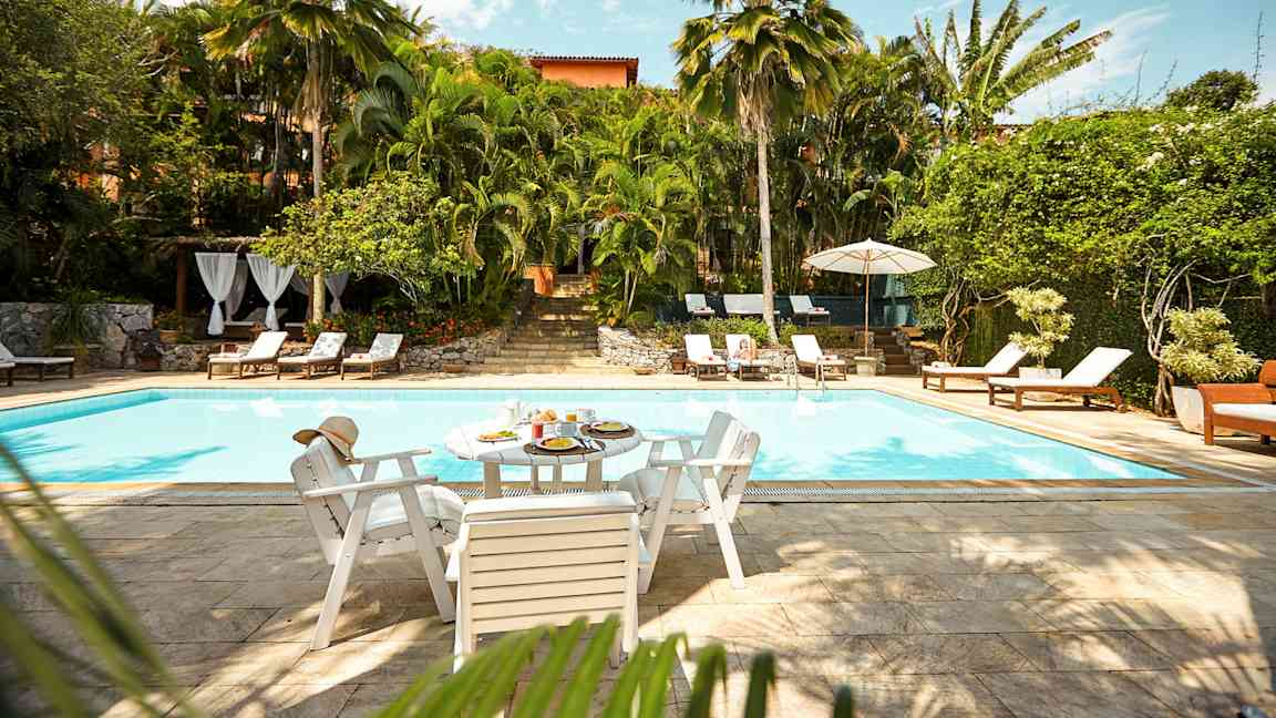 En udendørs pool omgivet af frodigt grønt og liggestole. Et bord med mad og drikke står ved poolkanten. (AI-genereret tekst)