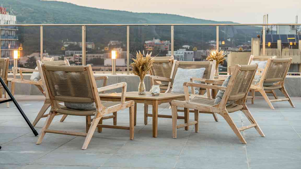 Terrasse med en gruppe træmøbler, herunder stole og bord. Udsigt over byen og grønne bakker i baggrunden. (AI-genereret tekst)