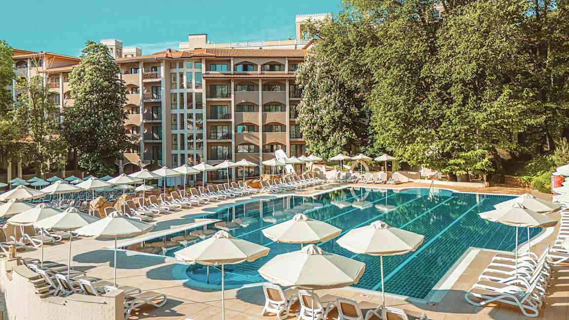 Poolområde med liggestole og parasoller, i baggrunden ses en hotelbygning og grønne træer. (AI-genereret tekst)