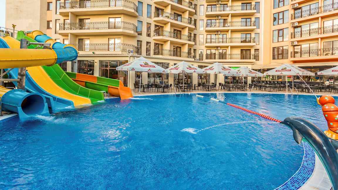 Billedet viser en swimmingpool med vandrutsjebaner i forskellige farver foran et hotel, med udemøbler og parasoller ved poolområdet. (AI-genereret tekst)