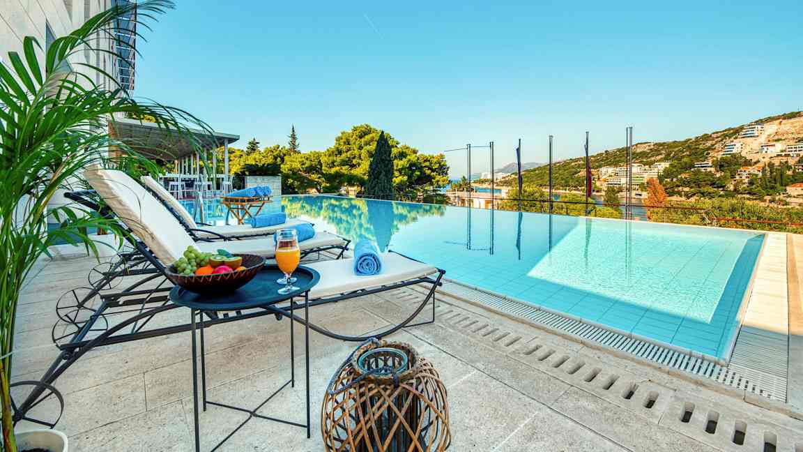 En udsigt over en infinitypool med liggestole, borde med frugt og drikke ved siden af, samt grønne omgivelser og himmel i baggrunden. (AI-genereret tekst)