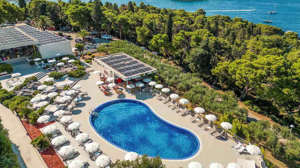 Udsigt over en udendørs pool omgivet af liggestole med parasoller, beliggende nær kysten med udsigt over skov og hav. (AI-genereret tekst)