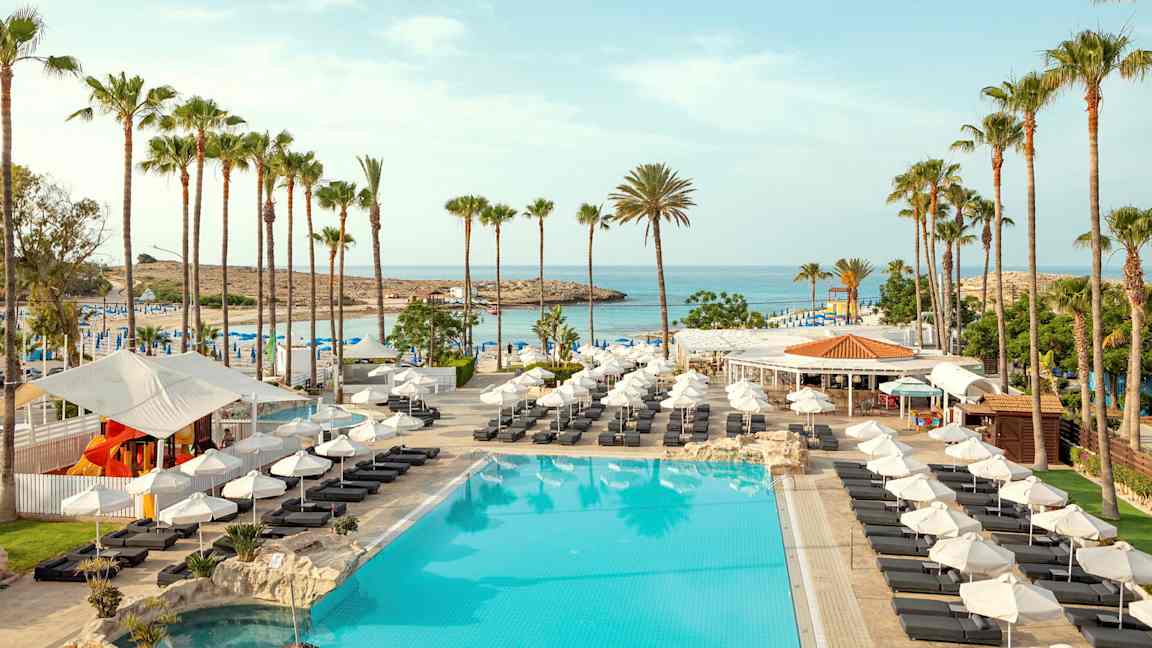 Billede af en pool med liggestole, parasoller og palmer ved en strand. (AI-genereret tekst)