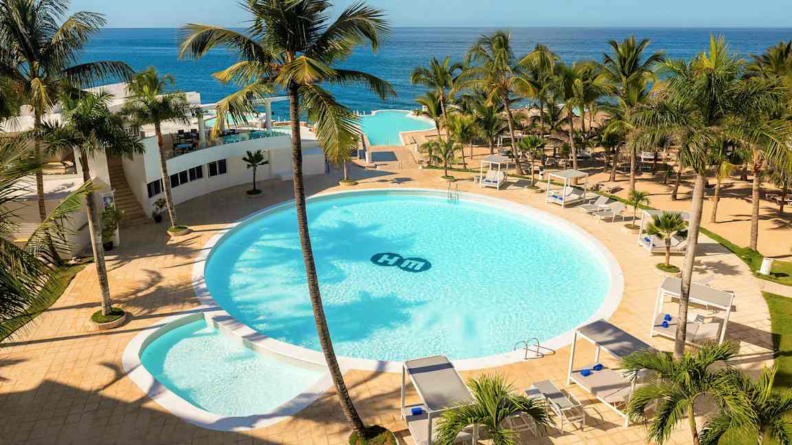 Et hotelområde med en stor rund pool omgivet af palmer, solsenge og cabanaer. Havudsigt i baggrunden. (AI-genereret tekst)