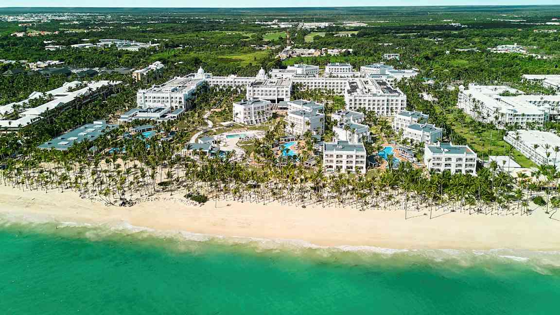 Luftfoto af et hotelkompleks ved en strand, inklusive poolområde, omgivet af grønne omgivelser. (AI-genereret tekst)