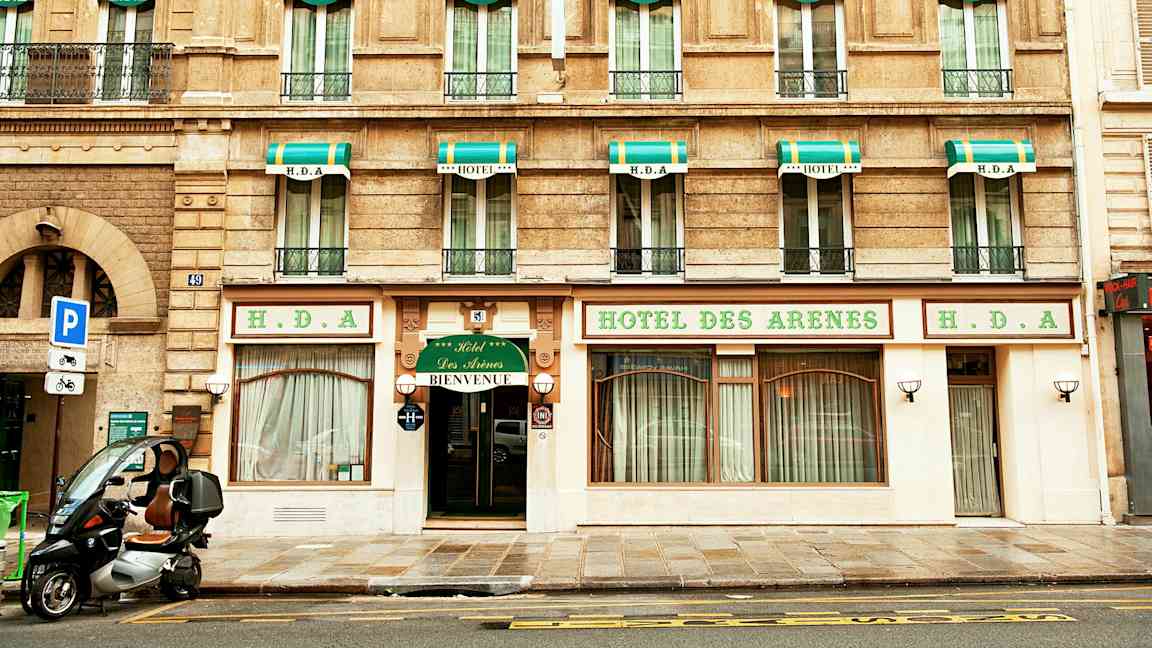 Bygning med hotellfacade, skilt med teksten 'Hotel des Arenes' og grønne og hvide markiser over vinduerne. (AI-genereret tekst)