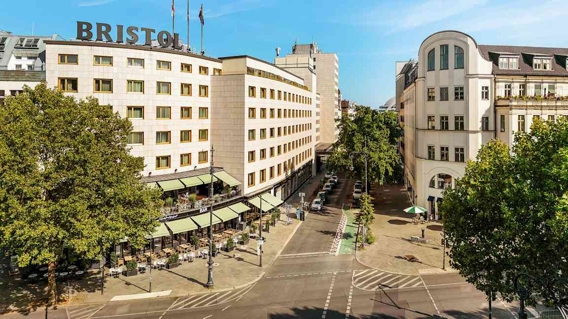Et bylandskab med en bygning med teksten "Bristol" på taget, omgivet af træer og en gade med udendørs servering. (AI-genereret tekst)