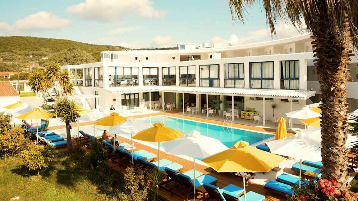 Et hotel med hvid facade, en udendørs pool omgivet af liggestole med gule og hvide parasoller samt beplantning i forgrunden. (AI-genereret tekst)