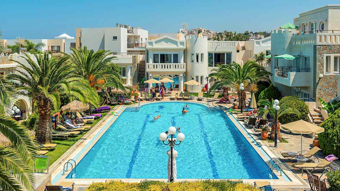 En pool omgivet af palmer, liggestole og bygninger under en klar blå himmel. (AI-genereret tekst)