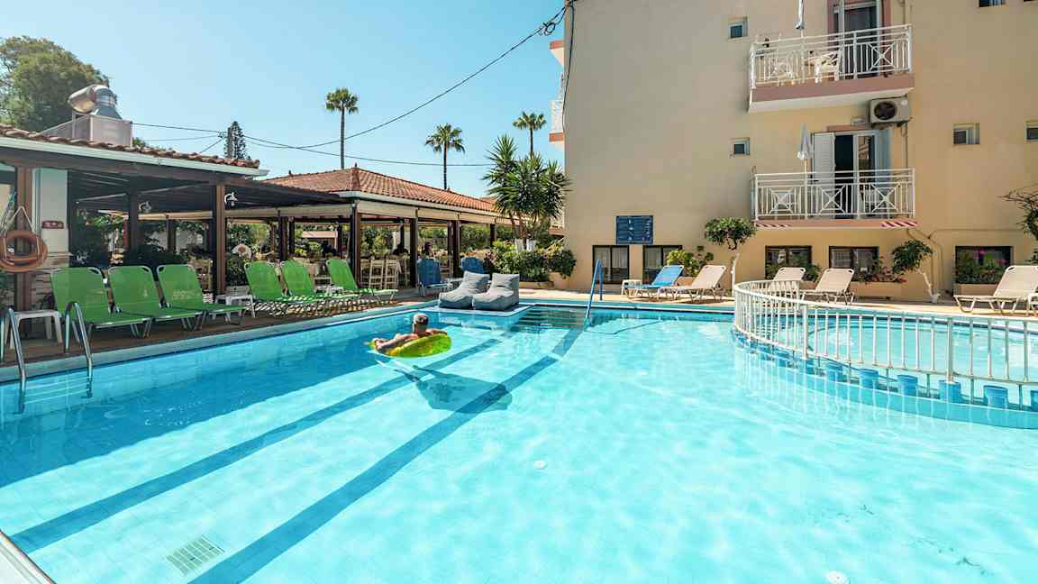 Hotel med liggestole omkring en udendørs pool og en person, der ligger på en oppustelig madras i vandet. (AI-genereret tekst)