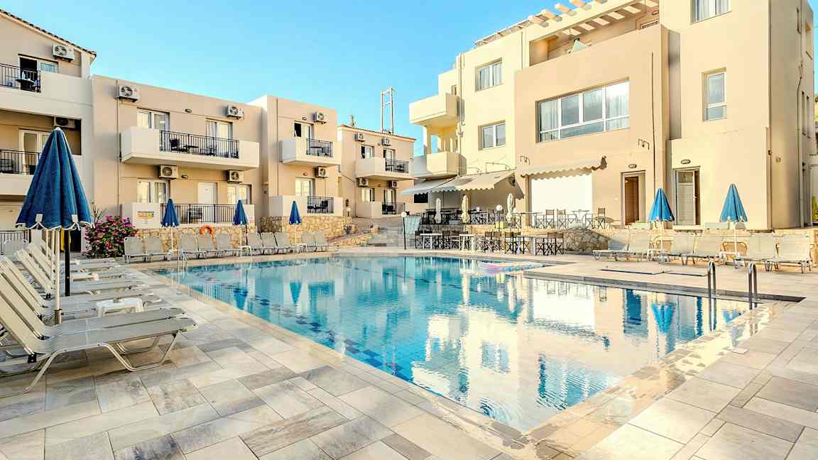 Udendørs swimmingpool omgivet af liggestole og parasoller foran en beige beboelsesbygning. (AI-genereret tekst)