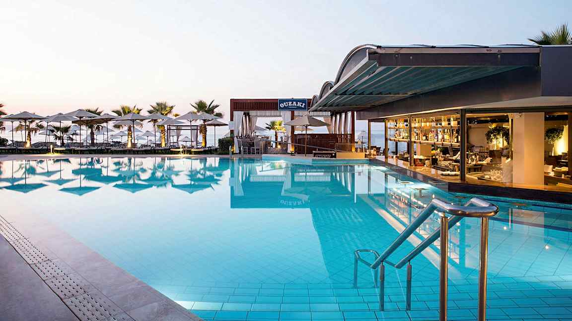 En stor pool ved en hotelrestaurant med udsigt over havet. Solstole og parasoller omgiver området. (AI-genereret tekst)