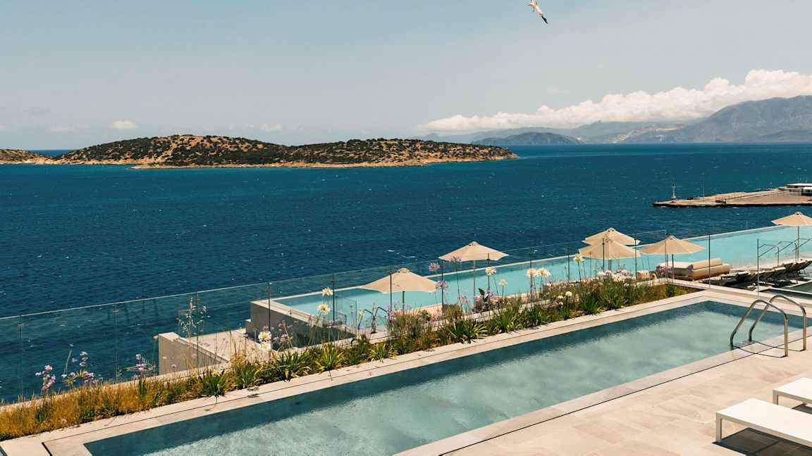 Billede af en infinitypool med udsigt over havet og omkringliggende øer. Et glasrækværk omgiver poolområdet, og parasoller er placeret ved poolkanten. (AI-genereret tekst)