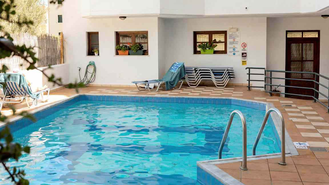 En lille swimmingpool omgivet af en terrasse med liggestole og en bygning med hvide vægge. (AI-genereret tekst)