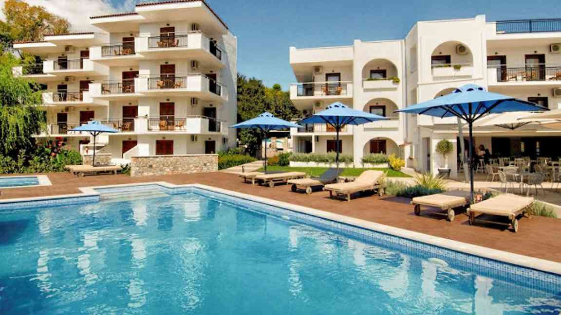 Hvid bygning med altaner, pool og liggestole med blå parasoller. (AI-genereret tekst)