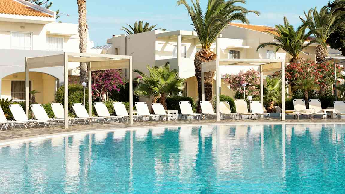 Poolområde med liggestole og parasoller omgivet af palmer og blomstrende buske foran lyse bygninger. (AI-genereret tekst)