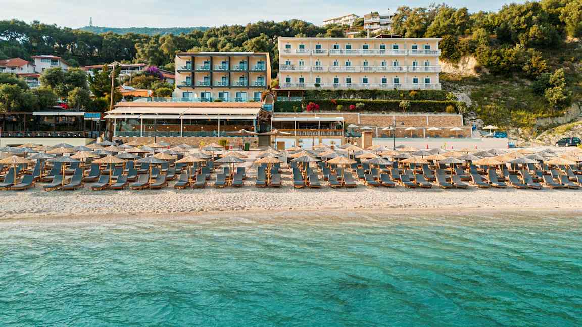 En sandstrand ved havet med liggestole og parasoller. I baggrunden ses hotelbygninger og beplantning. (AI-genereret tekst)