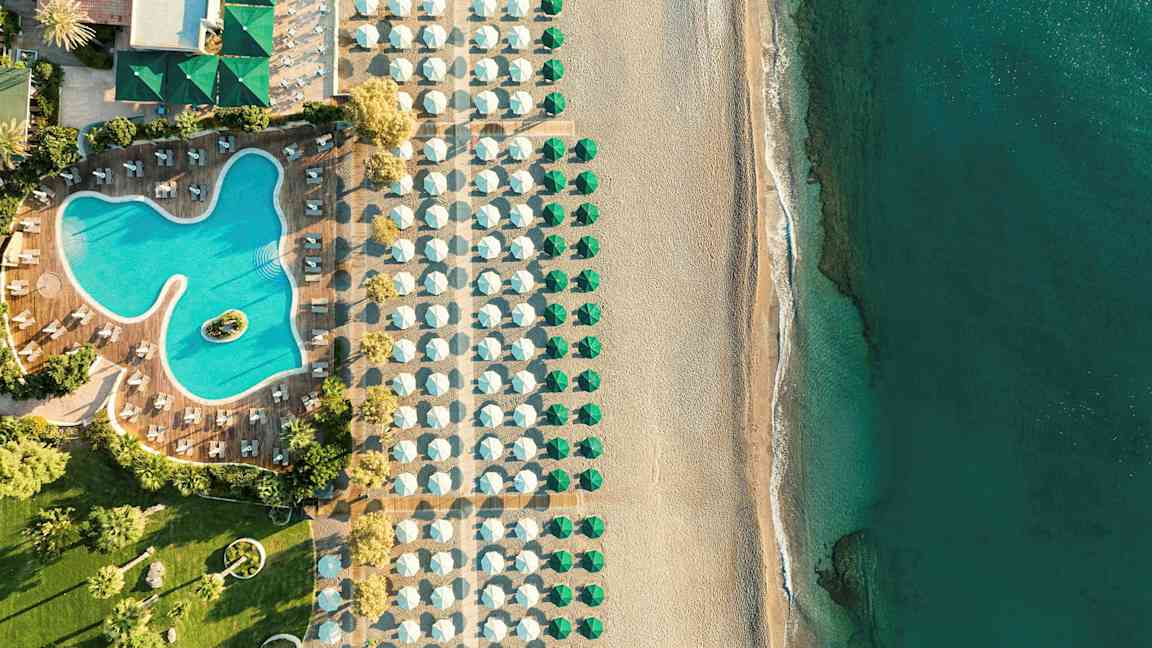 Luftfoto af en hotelpool med omkringliggende liggestole, ved siden af en strand med parasoller og hav. (AI-genereret tekst)