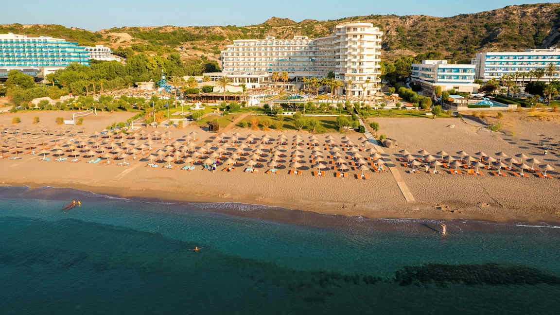 Et hotel ved stranden med liggestole og parasoller foran samt en klar blå himmel i baggrunden. (AI-genereret tekst)