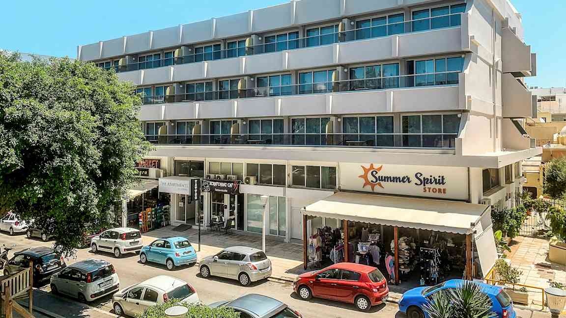 En moderne bygning med flere etager og butikker i stueetagen. Foran bygningen holder parkerede biler. (AI-genereret tekst)