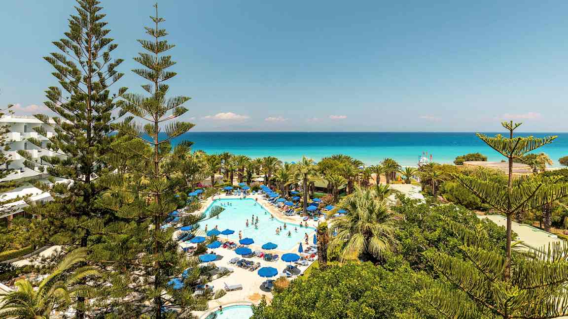 Billede af hotelområde med pool og solsenge omgivet af grønne områder og udsigt over havet. (AI-genereret tekst)