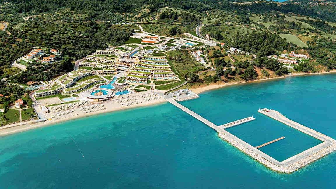 Luftfoto af et ferieområde med hoteller, swimmingpools og strand med liggestole, omgivet af grønne områder og hav. (AI-genereret tekst)