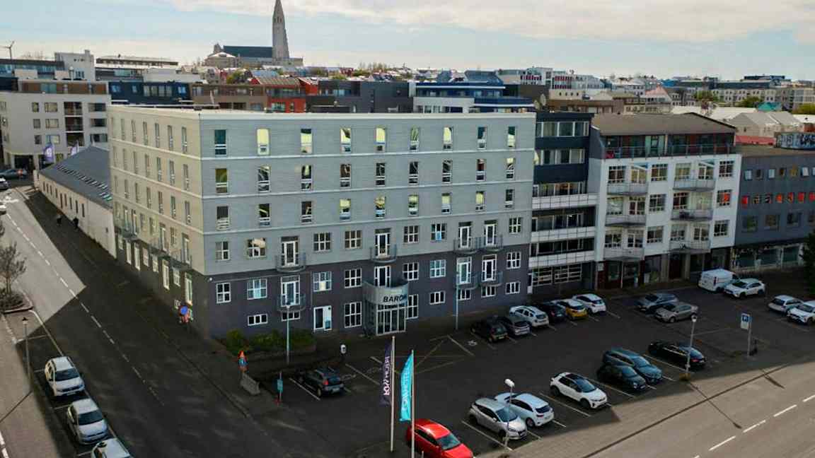 Flere moderne bygninger omgiver et åbent parkeringsområde, med biler parkeret på jorden og en kirke synlig i baggrunden. (AI-genereret tekst)