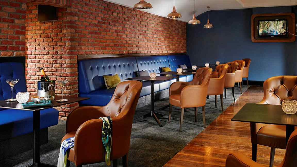 En restaurant eller lounge med blåpolstrede bænke og brune læderstole. Væggene er af røde mursten, og der er små borde med dekorative lys. (AI-genereret tekst)