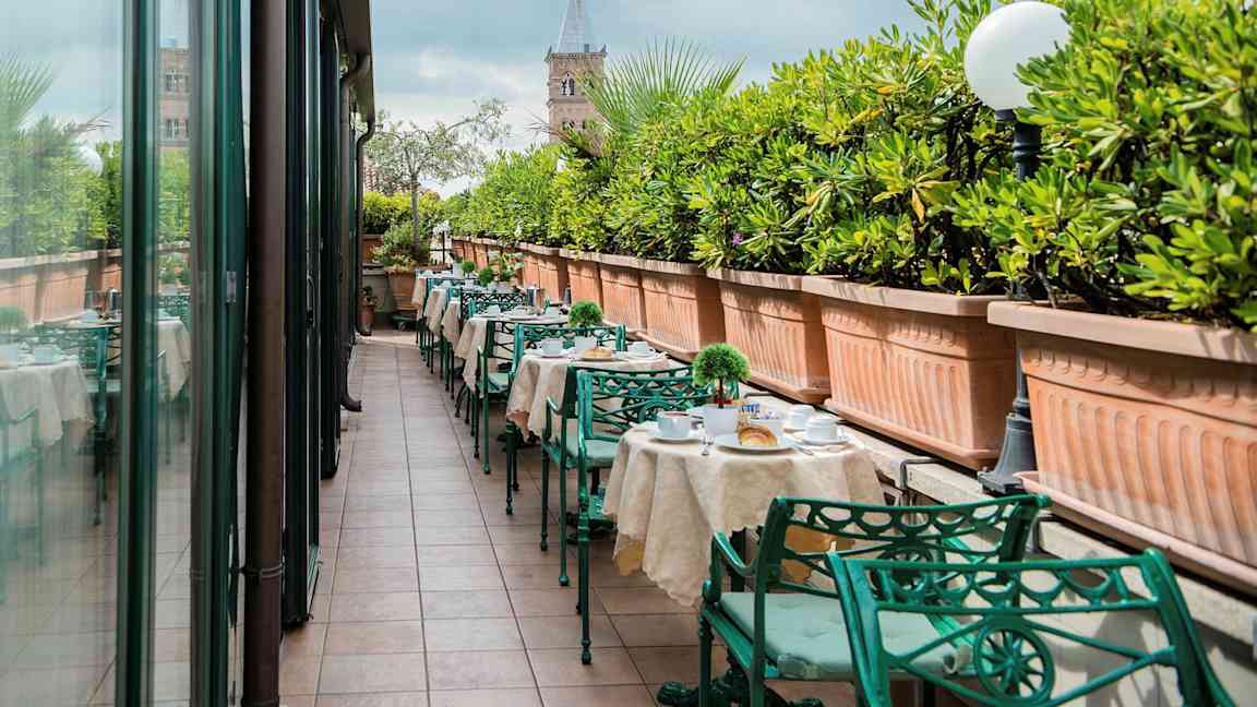 En udendørs terrasse med dækkede borde og grønne metalstole, kantet af store plantekasser med grønne planter. (AI-genereret tekst)