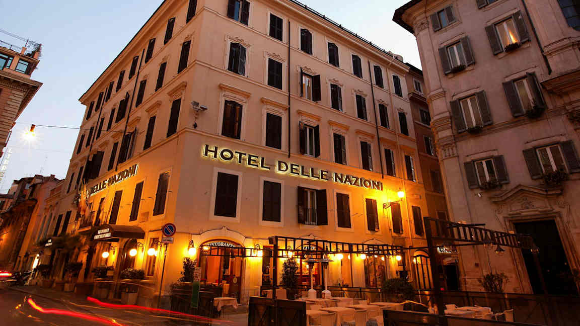 Billede af en bygning med skilt, der siger Hotel Delle Nazioni, oplyst med aftenbelysning. (AI-genereret tekst)