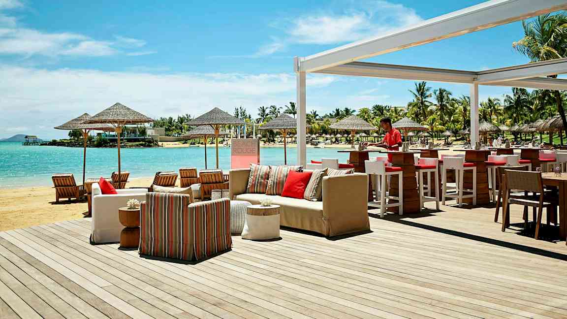 Udendørs serveringsområde ved stranden med bar og siddepladser, samt udsigt over havet og omkringliggende palmer. (AI-genereret tekst)