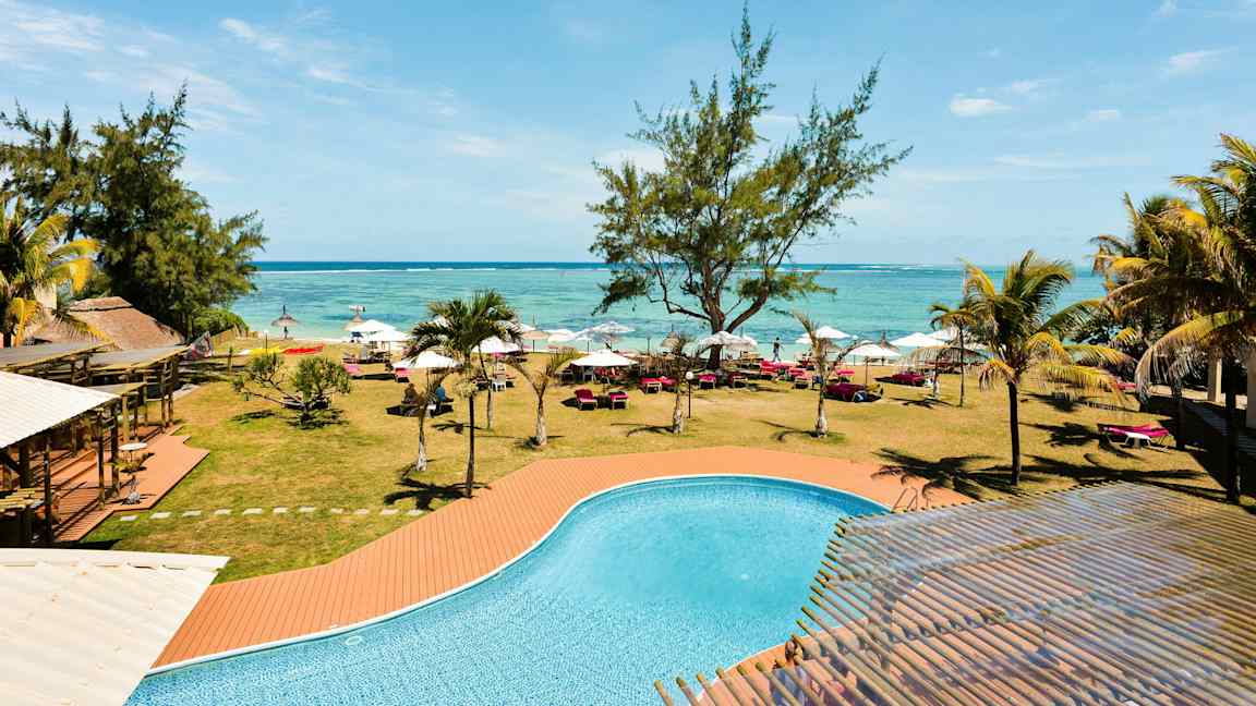 En pool med turkis vand foran en græsplæne med træer, liggestole og parasoller langs en strand. (AI-genereret tekst)