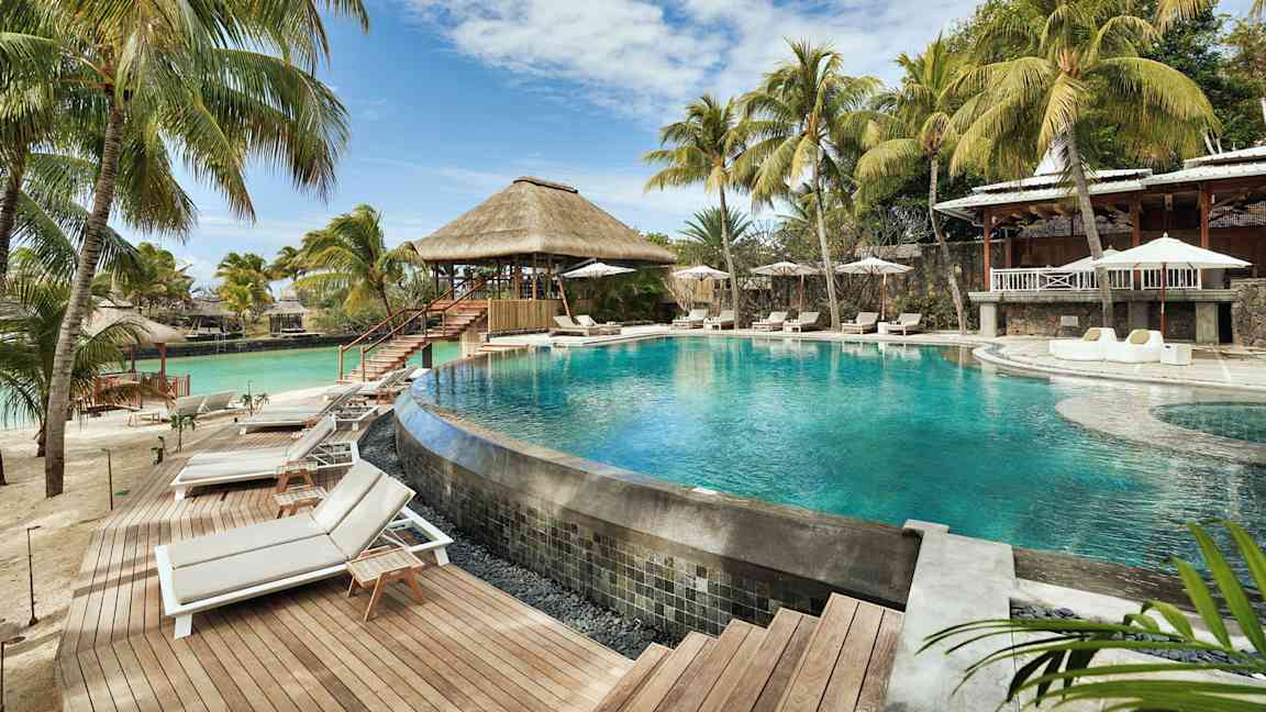 En pool omgivet af liggestole og parasoller, med palmer og en bygning med stråtag i baggrunden. (AI-genereret tekst)