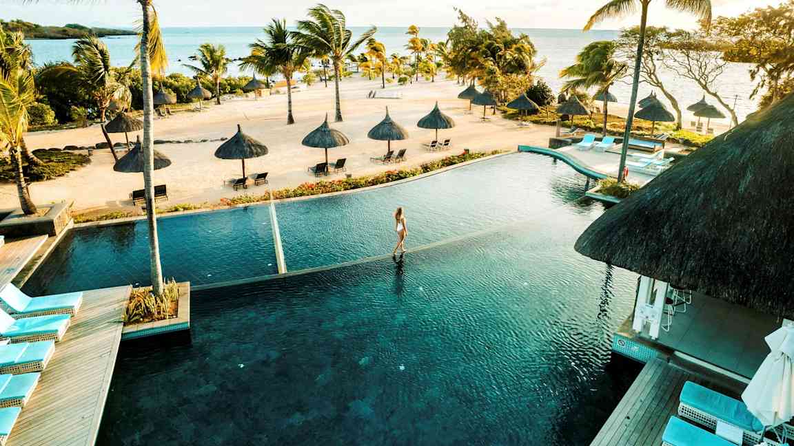 En tropisk strand med palmer og parasoller ved havet, en infinity-pool i forgrunden. (AI-genereret tekst)