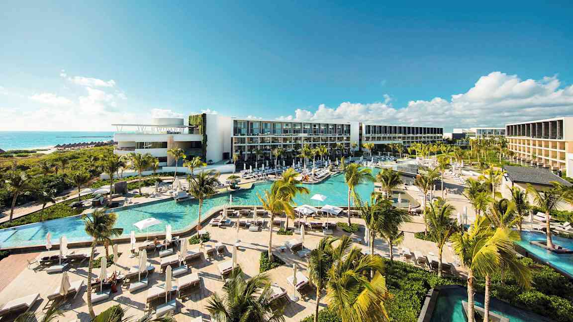 Et moderne hotelkompleks med flere pools, frodig vegetation og palmer, beliggende ved kysten med udsigt over havet under en klar blå himmel. (AI-genereret tekst)